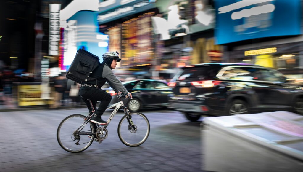 Bicycle courier with a large insulated backpack rides through Times Square traffic at night, captured with motion blur.